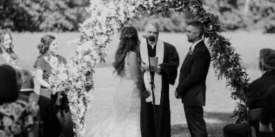 Black and white photograph of an outdoor wedding ceremony featuring a bride and groom standing under a floral arch with an officiant and seated guests.