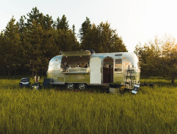 A silver Airstream trailer converted into a mobile bar with an open service window and glassware display, situated in a grassy field with pine trees in the background.