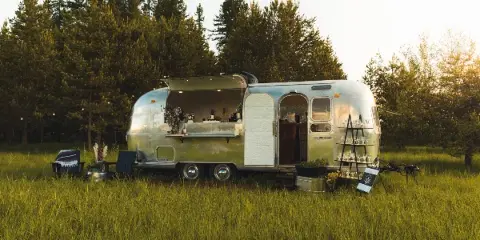 A silver Airstream trailer converted into a mobile bar with an open service window and glassware display, situated in a grassy field with pine trees in the background.