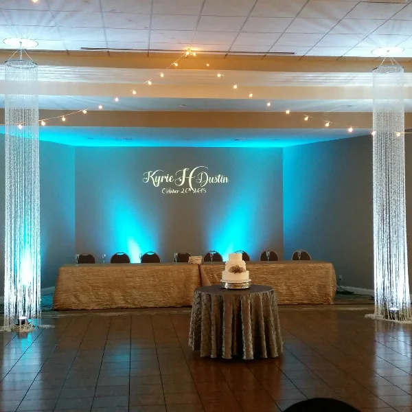 A wedding reception hall featuring a head table with a cake, illuminated by blue uplighting, string lights, and tall crystal columns.
