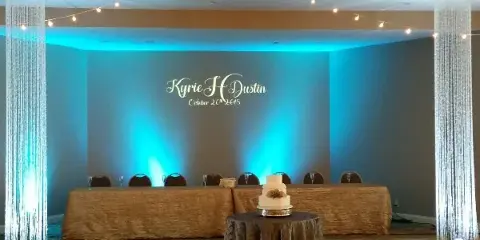 A wedding reception hall featuring a head table with a cake, illuminated by blue uplighting, string lights, and tall crystal columns.