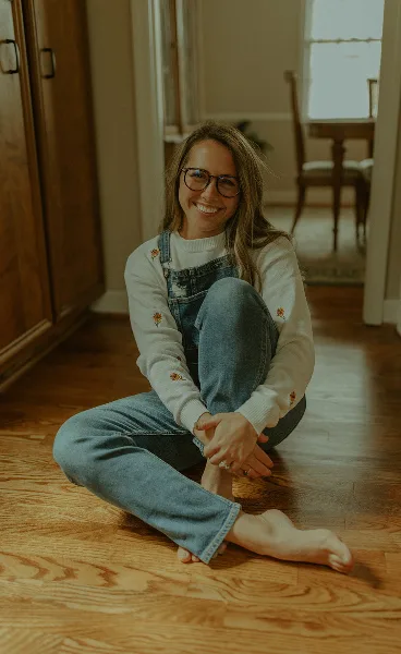 Portrait of a smiling woman with glasses and denim overalls sitting on a wooden floor.
