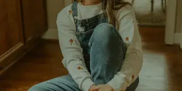 Portrait of a smiling woman with glasses and denim overalls sitting on a wooden floor.