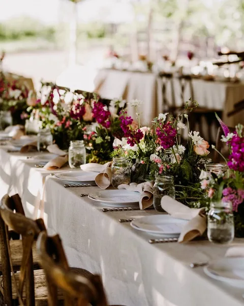 A long outdoor wedding table set with white linens, rustic wooden chairs, and lush floral centerpieces featuring purple and white blooms in mason jars.
