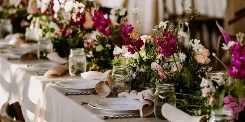 A long outdoor wedding table set with white linens, rustic wooden chairs, and lush floral centerpieces featuring purple and white blooms in mason jars.