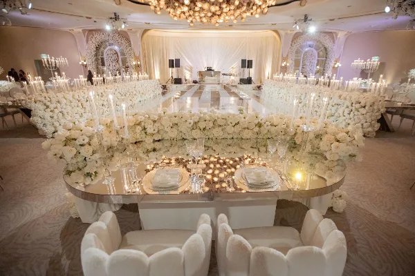 A long banquet table set with white linens, glassware, and floral arrangements, surrounded by white upholstered chairs in a decorated event hall.