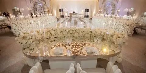 A long banquet table set with white linens, glassware, and floral arrangements, surrounded by white upholstered chairs in a decorated event hall.