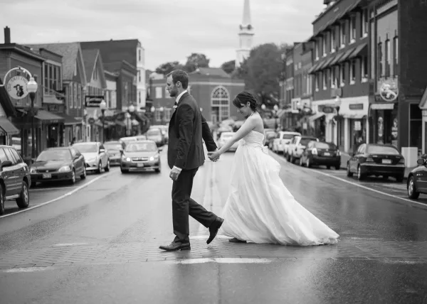 A black and white photograph of a bride and groom holding hands while walking down a wet street in a town.