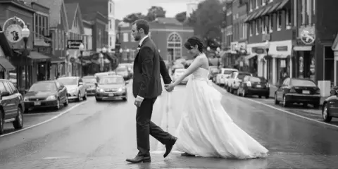 A black and white photograph of a bride and groom holding hands while walking down a wet street in a town.