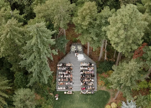 Aerial view of an outdoor wedding ceremony taking place on a wooden deck surrounded by tall evergreen trees.