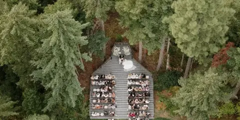 Aerial view of an outdoor wedding ceremony taking place on a wooden deck surrounded by tall evergreen trees.