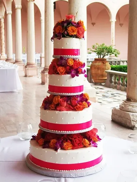 A four-tier white wedding cake decorated with vibrant orange and red roses and pink ribbons, displayed on a table in a venue with columns.