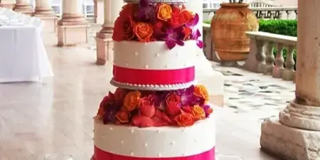 A four-tier white wedding cake decorated with vibrant orange and red roses and pink ribbons, displayed on a table in a venue with columns.