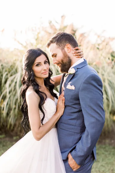 A bride with long, wavy dark hair and full makeup posing with a groom in an outdoor setting.