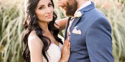 A bride with long, wavy dark hair and full makeup posing with a groom in an outdoor setting.