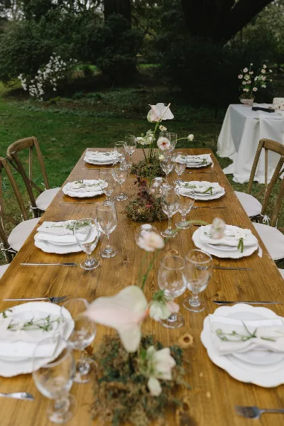 A rustic wooden farm table set with crossback chairs, white plates, and glassware in an outdoor garden setting.