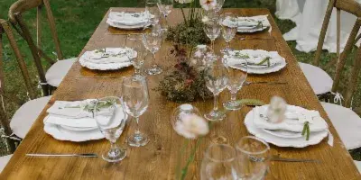 A rustic wooden farm table set with crossback chairs, white plates, and glassware in an outdoor garden setting.