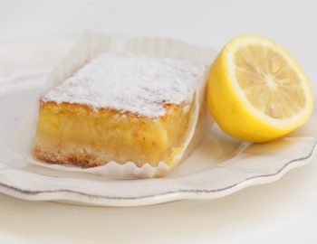 A square lemon bar dusted with powdered sugar served on a white plate next to a lemon wedge.