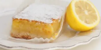 A square lemon bar dusted with powdered sugar served on a white plate next to a lemon wedge.