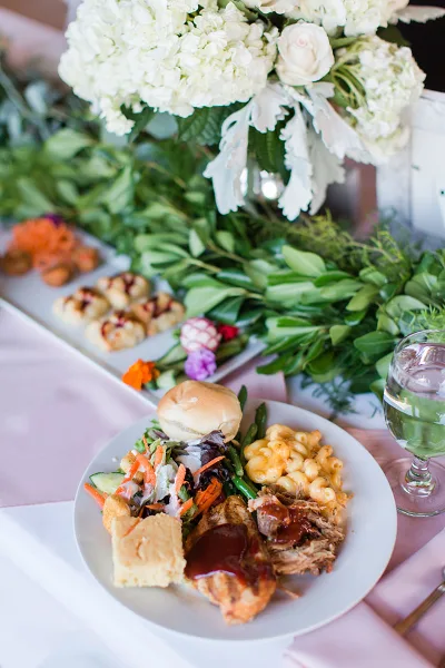 A plated catering dish with macaroni and cheese, pulled pork, and a side salad on a table with floral arrangements.