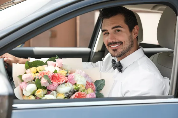 A smiling chauffeur in a white shirt and bow tie holding a large bouquet of flowers inside a car.