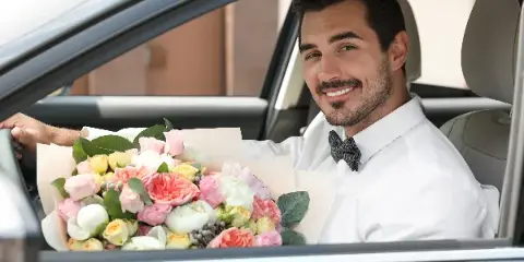 A smiling chauffeur in a white shirt and bow tie holding a large bouquet of flowers inside a car.