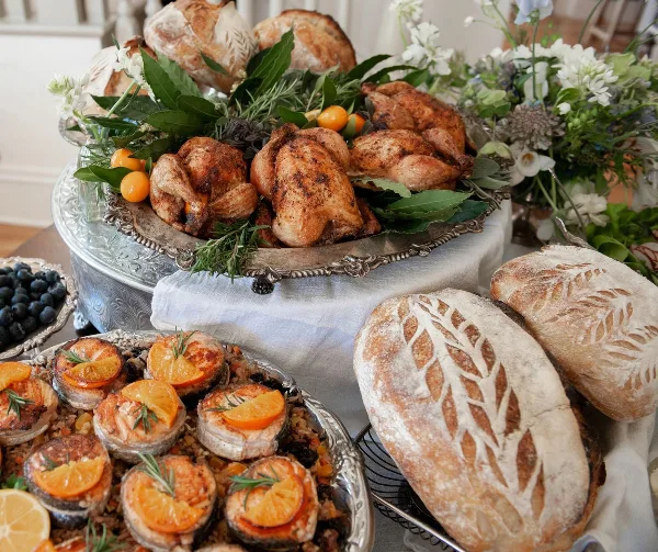 A catering table display featuring roasted chickens, artisan bread loaves, and fresh floral arrangements.