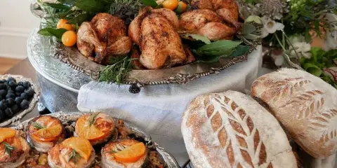 A catering table display featuring roasted chickens, artisan bread loaves, and fresh floral arrangements.