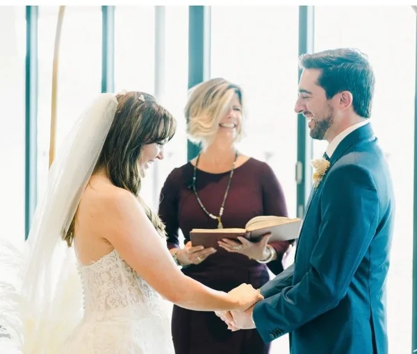 A wedding officiant stands between a bride and groom during a ceremony, holding an open book while the couple holds hands.