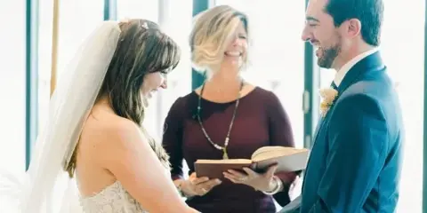 A wedding officiant stands between a bride and groom during a ceremony, holding an open book while the couple holds hands.