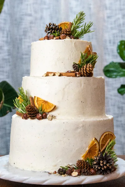Three-tier white wedding cake decorated with pinecones, rosemary sprigs, dried orange slices, and nuts on a white cake stand.