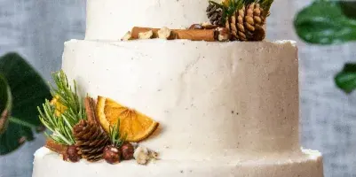 Three-tier white wedding cake decorated with pinecones, rosemary sprigs, dried orange slices, and nuts on a white cake stand.