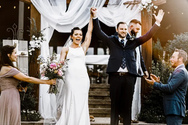 A bride and groom raising their hands in celebration at a wedding ceremony, surrounded by guests and floral decorations.
