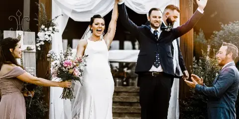 A bride and groom raising their hands in celebration at a wedding ceremony, surrounded by guests and floral decorations.