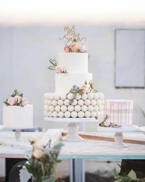 A three-tier white wedding cake decorated with pink roses and a gold 'Mr & Mrs Smith' topper, displayed on a cake stand alongside two smaller cakes.