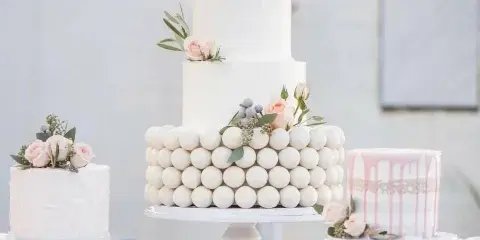 A three-tier white wedding cake decorated with pink roses and a gold 'Mr & Mrs Smith' topper, displayed on a cake stand alongside two smaller cakes.