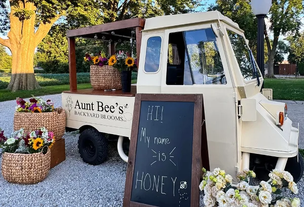 A cream-colored mobile flower truck branded 'Aunt Bee's Backyard Blooms' parked outdoors, displaying baskets of sunflowers and mixed blooms, with a chalkboard sign reading 'HI! My name is HONEY'.