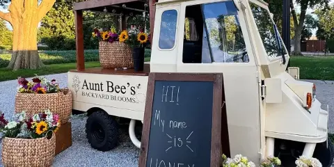 A cream-colored mobile flower truck branded 'Aunt Bee's Backyard Blooms' parked outdoors, displaying baskets of sunflowers and mixed blooms, with a chalkboard sign reading 'HI! My name is HONEY'.