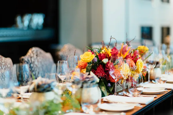 A close-up view of an elegant event table setting featuring a vibrant floral centerpiece with yellow and red flowers, glassware, and white plates.