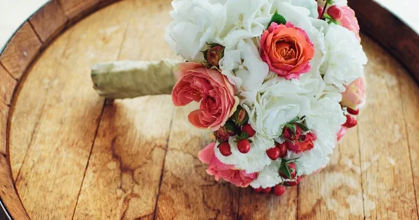 A hand-tied wedding bouquet with white hydrangeas, pink and orange roses, and red berries resting on a wooden barrel top.