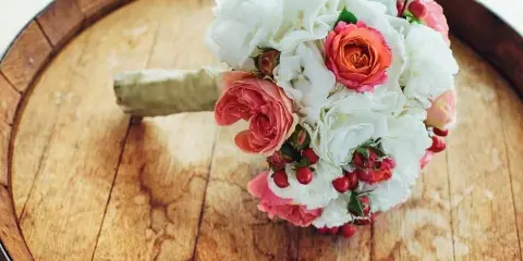 A hand-tied wedding bouquet with white hydrangeas, pink and orange roses, and red berries resting on a wooden barrel top.