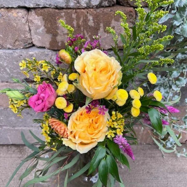 A mixed bouquet of yellow and pink blooms in a clear glass vase against a stone wall