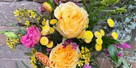 A mixed bouquet of yellow and pink blooms in a clear glass vase against a stone wall