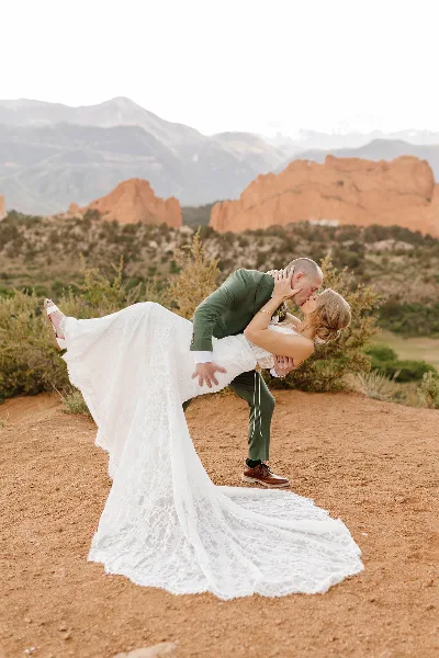 A bride in a white lace gown and a groom in a green suit kissing during a dip pose in a red rock landscape.