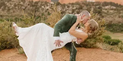 A bride in a white lace gown and a groom in a green suit kissing during a dip pose in a red rock landscape.