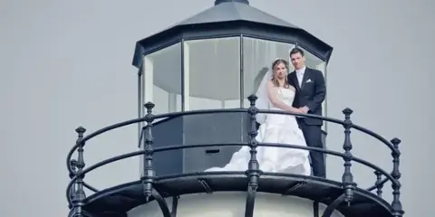 A bride and groom standing on the balcony of a lighthouse.
