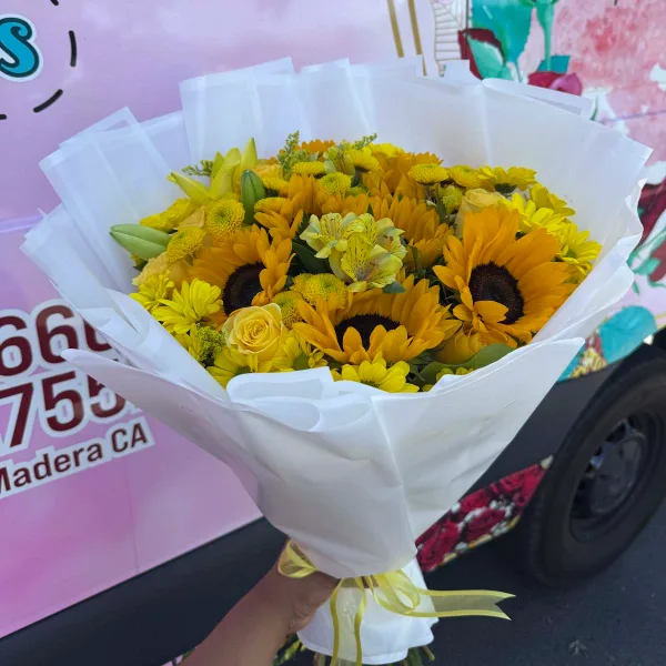 A hand holding a bouquet of yellow sunflowers and roses wrapped in white paper, positioned in front of a delivery van with Madera, CA text.