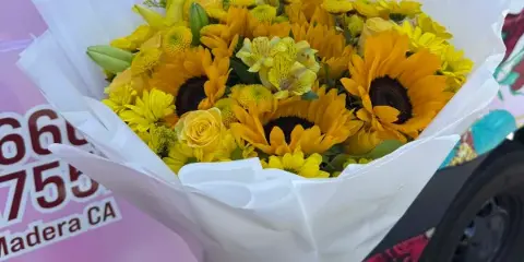 A hand holding a bouquet of yellow sunflowers and roses wrapped in white paper, positioned in front of a delivery van with Madera, CA text.