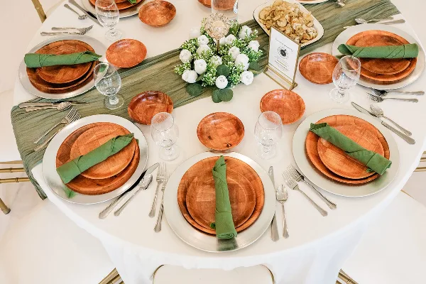 Styled catering table with wooden plates, green napkins, glassware, white floral centerpiece, and a dish of food.
