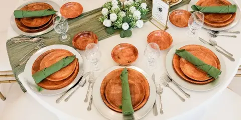 Styled catering table with wooden plates, green napkins, glassware, white floral centerpiece, and a dish of food.